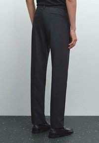 Personne portant un pantalon noir sur mesure et des mocassins noirs, se tenant contre un fond gris clair uni.