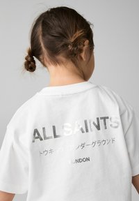 Balta medvilninė marškinėlių su trumpomis rankovėmis, su laisvu siluetu, ant nugaros yra sidabrinės spalvos metalinis užrašas "ALLSAINTS" ir "LONDON".
