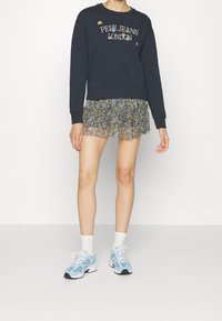 Felpa navy con il testo "Pepe Jeans London", gonna a strati floreale e scarpe da ginnastica azzurro chiaro, abbinate a calzini bianchi e un fit moderno.
