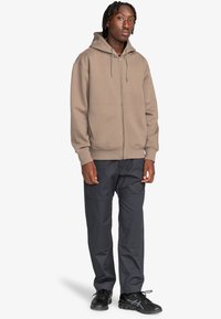Light brown zip-up hoodie with drawstring hood, worn with dark gray pants and black athletic shoes. Textured fabric, relaxed fit.