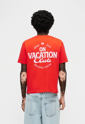 Person mit dunklem lockigem Haar und Vitiligo-Flecken auf den Armen, trägt ein rotes T-Shirt mit der Aufschrift "On Vacation Club" und hellblaue Jeans, vom Betrachter abgewandt.