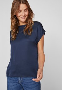Mørkeblå bluse med korte ermer laget av glatt stoff, med rund hals og løs passform, sammen med blå denimjeans.