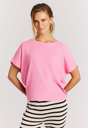 Vrouw draagt een loszittend roze shirt met korte mouwen en zwart-wit horizontaal gestreepte broek, staand met handen in de zij.