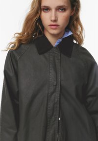 Giacca nera con colletto in velluto a coste, chiusura con zip e vestibilità leggermente rilassata. Presenta maniche raglan e una texture liscia.