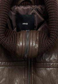 Giacca in pelle marrone con colletto a coste. Presenta una zip argentata e un'etichetta in tessuto con la scritta "MNG". Finitura in pelle testurizzata.