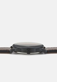 Runde schwarze Uhr mit strukturiertem Gehäuse und silbernen Akzenten. Der braune Lederarmband erstreckt sich vom Gehäuse und zeigt ein minimalistisches Design.