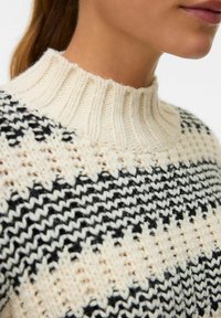 Megzti svarkas su aukšta, raštuota kremine spalva apykakle. Kūnas puikuojasi juoda ir kremine spalva geometriškais raštais su įvairiomis tekstūromis.