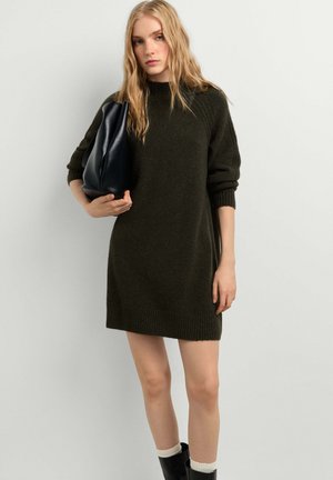 Blonde Frau in einem dunkelgrünen Strickkleid mit einer schwarzen Lederhandtasche, stehend vor einem einfarbigen hellen Hintergrund.