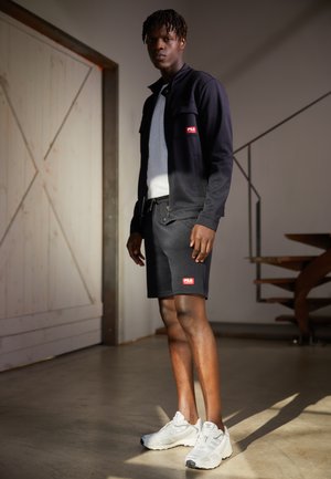 Giacca nera con zip e pantaloni corti abbinati, entrambi con toppe rosse con il marchio; sneakers bianche testurizzate completano l'outfit. Sfondo neutro.