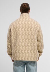 Beige fleece takki, jossa on korkea kaulus, vinkkakuosi ja pehmeä pinta, käytettävä vaaleansinisten farkkujen kanssa.
