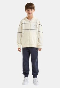 Felpa con cappuccio color crema con cerniera e strisce nere, arricchita da un dettaglio logo. Abbinata a pantaloni della tuta blu navy e sneakers bianche con accenti blu.