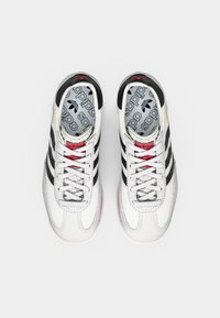 Fehér atlétikai edzőcipők három fekete csíkkal, ezüst részletekkel és piros fűzőkkel. Texturált felsőrész és párnázott szegély. Nyomtatott logó a talpbetéten.