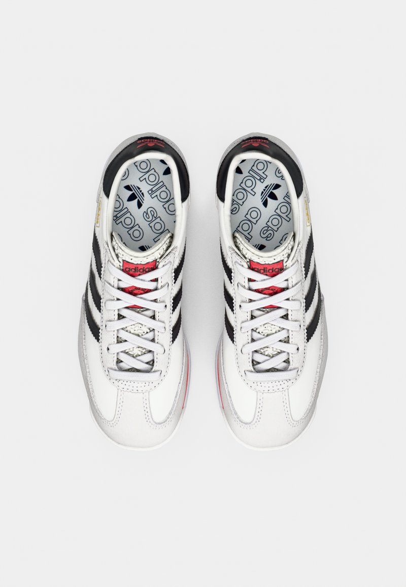 Baskets de sport blanches avec trois bandes noires, accents argentés et lacets rouges. Tige texturée et col rembourré. Logo imprimé sur les semelles intérieures.