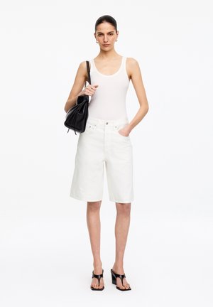 Femme portant un haut blanc sans manches, un short blanc longueur genou, des sandales à talons noires et portant un sac à épaule noir, debout sur fond blanc.