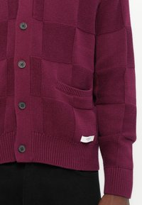 Cardigan lavorato a maglia color bordeaux con motivo a quadri, bottoni neri, tasca frontale e polsini a coste. Piccola etichetta del marchio cucita all'orlo.