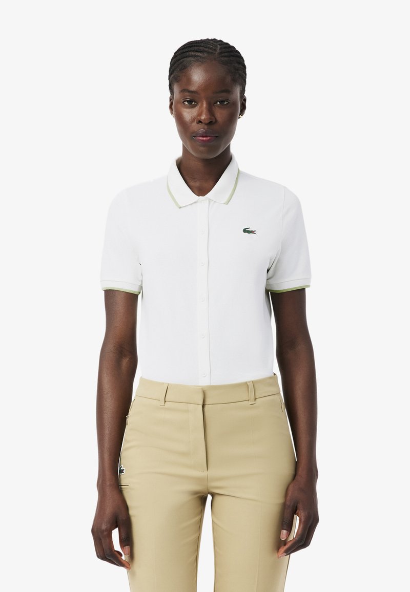 Frau mit geflochtenem Haar, die ein weißes Polohemd mit kurzen Ärmeln und beige Hose mit hoher Taille trägt, vor einem schlichten weißen Hintergrund stehend.