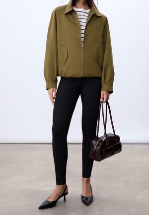 Donna che indossa una giacca verde oliva con zip, pantaloni neri slim, scarpe nere con tacco e cinturino posteriore, tiene in mano una borsa marrone scuro con due manici.
