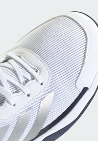 Weißer Sportschuh mit strukturiertem Mesh-Obermaterial, kontrastierenden silbernen Streifen und einer weichen Sohle mit dunklen Gummielementen.