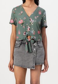Cropped top floreale verde con maniche corte e dettagli da annodare sul davanti, abbinato a una gonna in denim grigia a vita alta con design con bottoni e orlo sfilacciato.