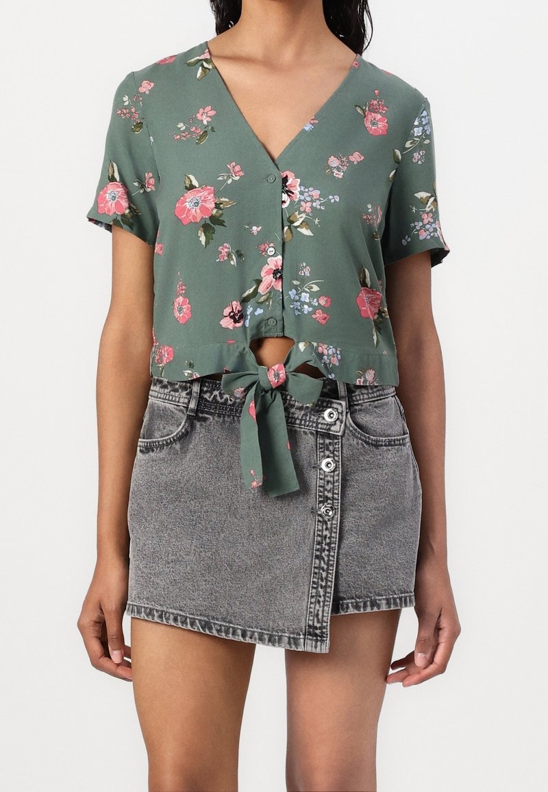 Cropped top floreale verde con maniche corte e dettagli da annodare sul davanti, abbinato a una gonna in denim grigia a vita alta con design con bottoni e orlo sfilacciato.