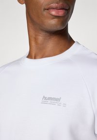 Zbliżenie osoby noszącej biały sweatshirt hummel z logo i tekstem na górnej części klatki piersiowej, neutralne szare tło.