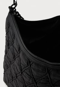 Borsa nera trapuntata con una morbida texture in tessuto, dotata di apertura con cerniera e una robusta tracolla a catena. Interno scuro e semplice.