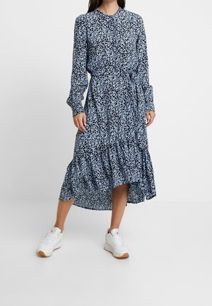 Kobieta w długiej, granatowej sukience w kwiaty z wiązanym w talii paskiem i białymi trampkami, stojąca na jasnym, jednolitym tle.