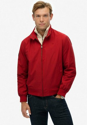 Man in een rode ritsjack over een beige ritstrui, met donkere jeans, staand tegen een witte achtergrond.