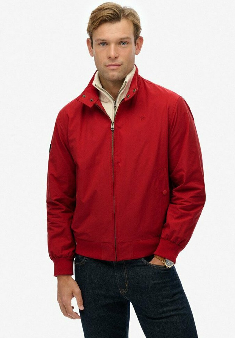 Man in een rode ritsjack over een beige ritstrui, met donkere jeans, staand tegen een witte achtergrond.