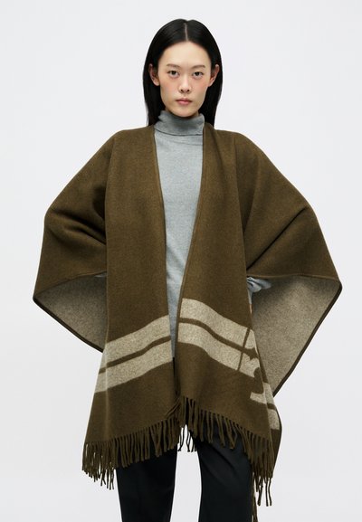 Poncho verde-azeitona com riscas cinza claro, bordas frisadas, material de lã macia e um design solto e fluido. Usado por cima de uma camisola de gola alta cinza.