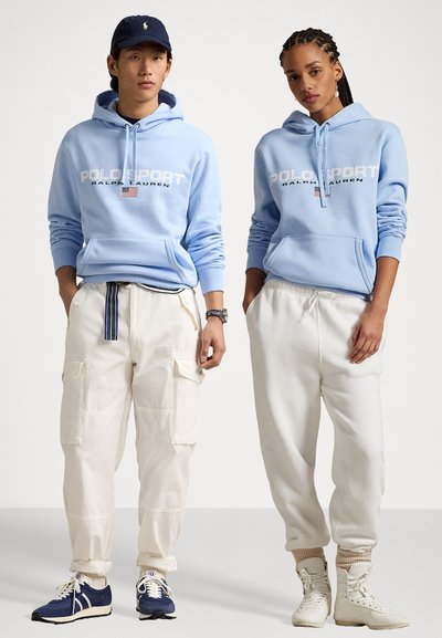 Ανοιχτό μπλε φούτερ με λογότυπο "POLOSPORT", κρέμα παντελόνι φόρμας και αθλητικά παπούτσια. Ο αρσενικός μοντέλο φοράει ναυτικό καπέλο; η θηλυκή μοντέλο έχει πλεξούδες.