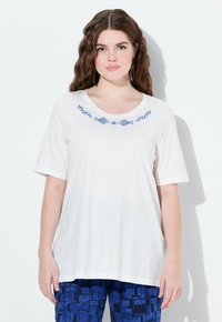 Bílá krátkosrstá tričko s výstřihem do V a modrou výšivkou ryb a listů na hrudi, nošené s vzorovanými modrými kalhotami.