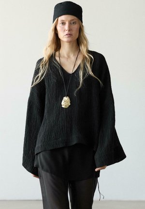 Žena v volném černém texturovaném topu, černých kalhotách, černé čelence a s velkým zlatým přívěskem na krku na jednobarevném pozadí.