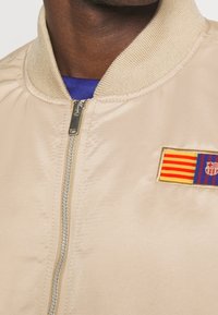 Beige Bomberjacke mit geripptem Kragen und Reißverschluss. Verfügt über ein gesticktes Barcelona-Flaggen-Patch auf der linken Brust.