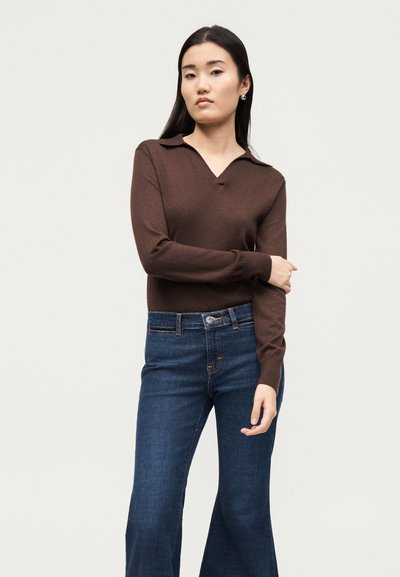 Γυναίκα που φοράει ένα καφέ πουλόβερ με μακριά μανίκια και γιακά και μπλε τζιν με ψηλή μέση, στέκεται με το ένα χέρι σταυρωμένο μπροστά στο σώμα της.