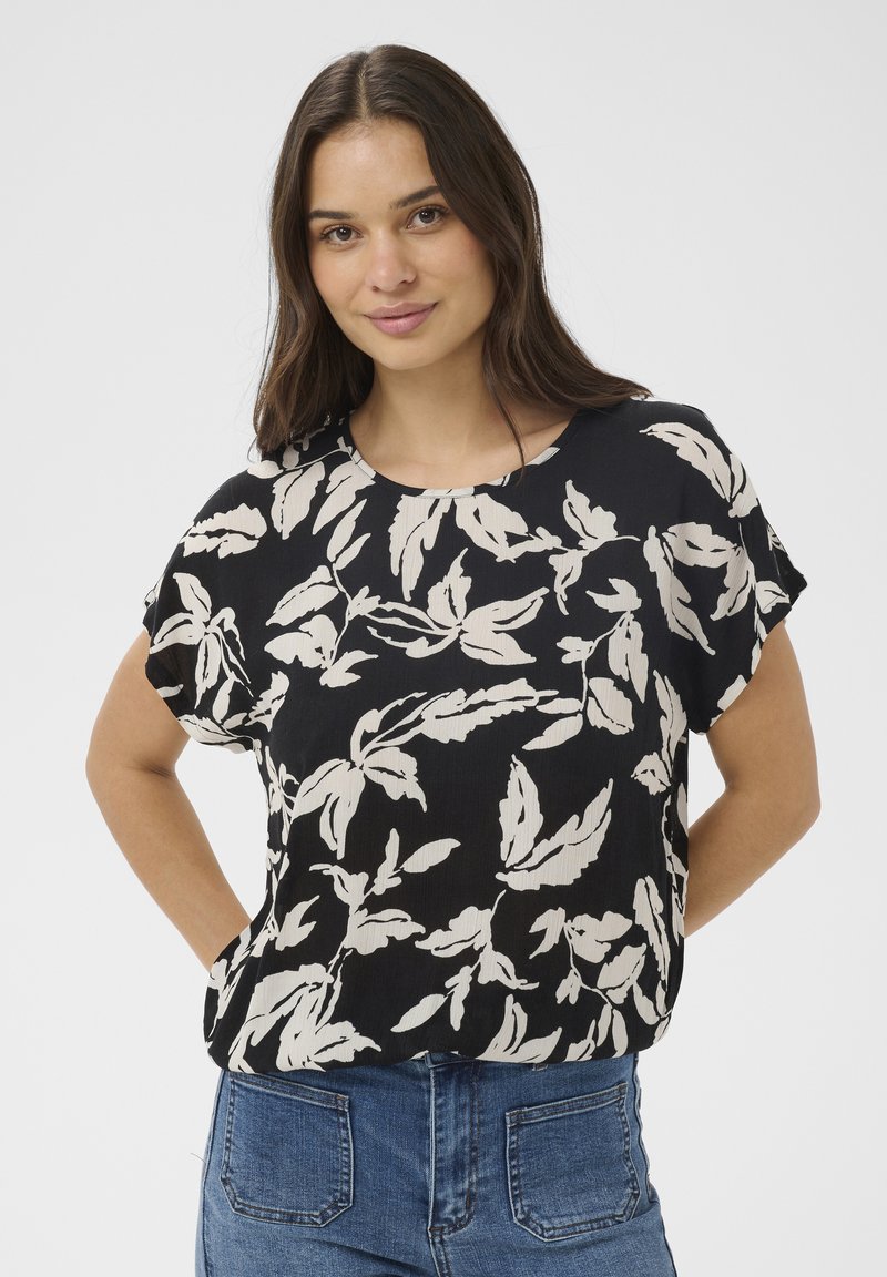 Jeune femme aux longs cheveux bruns portant un chemisier noir à motifs de feuilles beiges et un jean bleu, debout les mains derrière le dos.