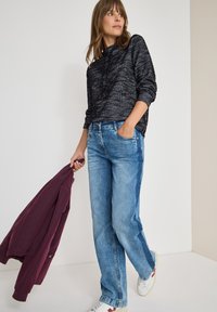Felpa nera con texture, jeans blu e sneakers bianchi. Modello tiene una giacca bordeaux, in piedi contro uno sfondo neutro.