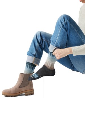 Person in blauer Jeans zieht einen gemusterten Socken an, neben ihr liegt ein hellbrauner Wildlederstiefel auf weißem Hintergrund.
