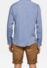 Světle modrá košile na knoflíky s dlouhými rukávy, doplněná béžovými kraťasy s ohrnutými nohavicemi. Oba kousky mají ležérní, volný střih.