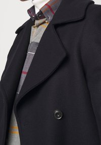 Schwarzer Wollmantel mit breitem Kragen und doppelter Knopfleiste. Darunter ein graues, kariertes Hemd mit roten und gelben Streifen.