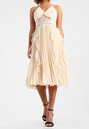 Kobieta ubrana w kremową plisowaną sukienkę midi z koronkowymi wykończeniami, z dekoltem halter i czarnymi sandałkami na kostkę, stojąca na jednolitym tle.