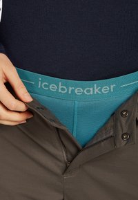 Teal midja på Icebreaker under grå byxor, vilket visar tygtextur och färgkontrast. Innehåller dragkedjor, knappar och en närbild på handens placering.