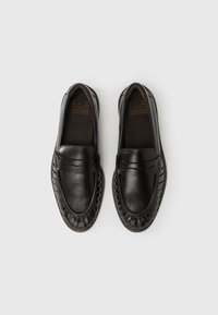 Μαύρα δερμάτινα μοκασίνια με λεία επεξεργασία, διακοσμητικό ράψιμο και κυματιστά άκρα σόλας, με ανυψωμένη μπροστινή πλάκα για εύκολη εφαρμογή κατά την εισαγωγή των ποδιών.