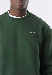 Uomo che indossa una felpa di colore verde scuro con la scritta "MILLION" in bianco sulla parte sinistra del petto, sopra una maglietta bianca. Sfondo bianco semplice.