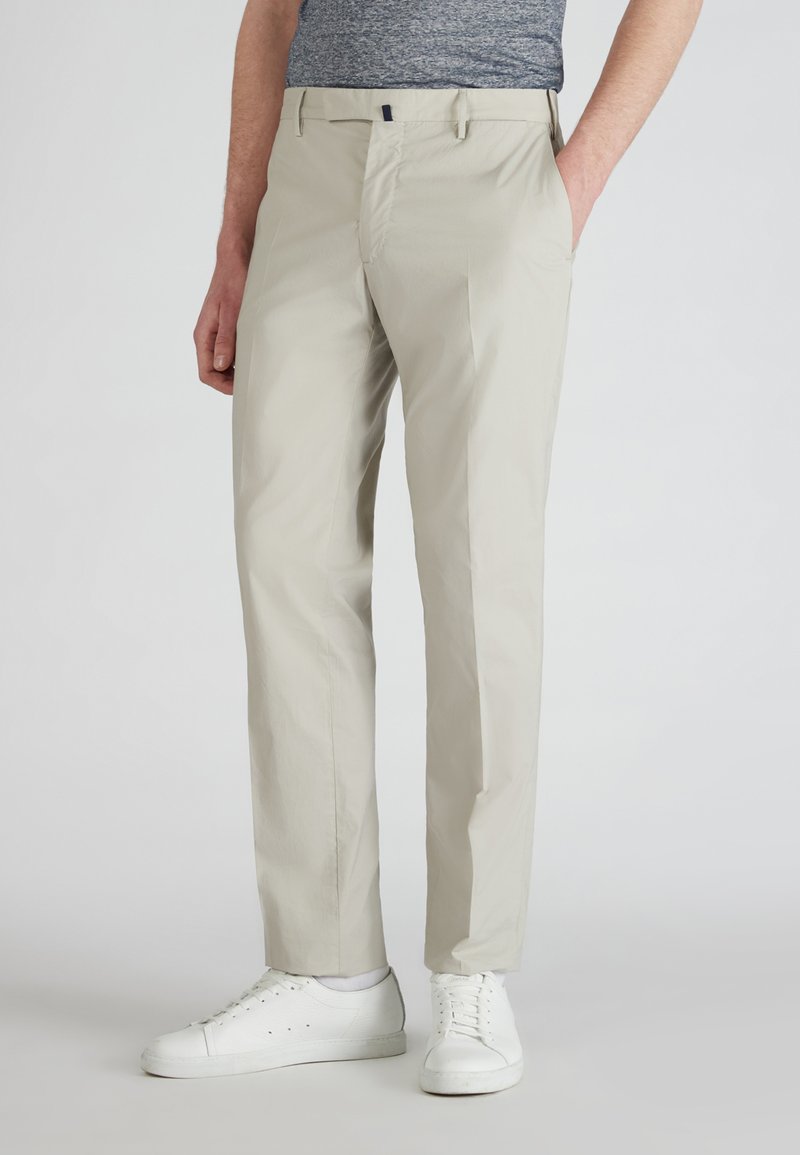 Homme portant un pantalon beige à jambe droite et des baskets blanches, se tenant avec une main dans la poche contre un fond léger uni.