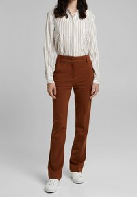 Hnědé kalhoty rovného střihu s knoflíkovým zapínáním, doplněné bílou pruhovanou košilí. Model má na sobě bílé tenisky a stojí na neutrálním pozadí.