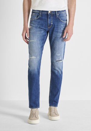 Hombre con vaqueros ajustados azules desgastados y zapatillas beige, de pie frente a un fondo blanco liso.