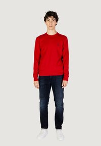 Maglione rosso a maniche lunghe con scollo rotondo, abbinato a jeans scuri e sneaker bianche. Presenta un piccolo logo sul petto.
