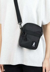 Mustat nylon-olkalaukku, jossa on etupaneelin vetoketjullinen tasku, säädettävä hihna ja logopaketti. Kompakti suorakaiteen muotoinen, sileä pinta.