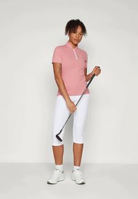 Ροζ γκολφ πόλο με ζιπ-κολάρο, συνδυασμένο με λευκά παντελόνια capri. Το μοντέλο κρατά ένα γκολφ κλαμπ, ενώ τα επιβλητικά αθλητικά παπούτσια ολοκληρώνουν την εμφάνιση.